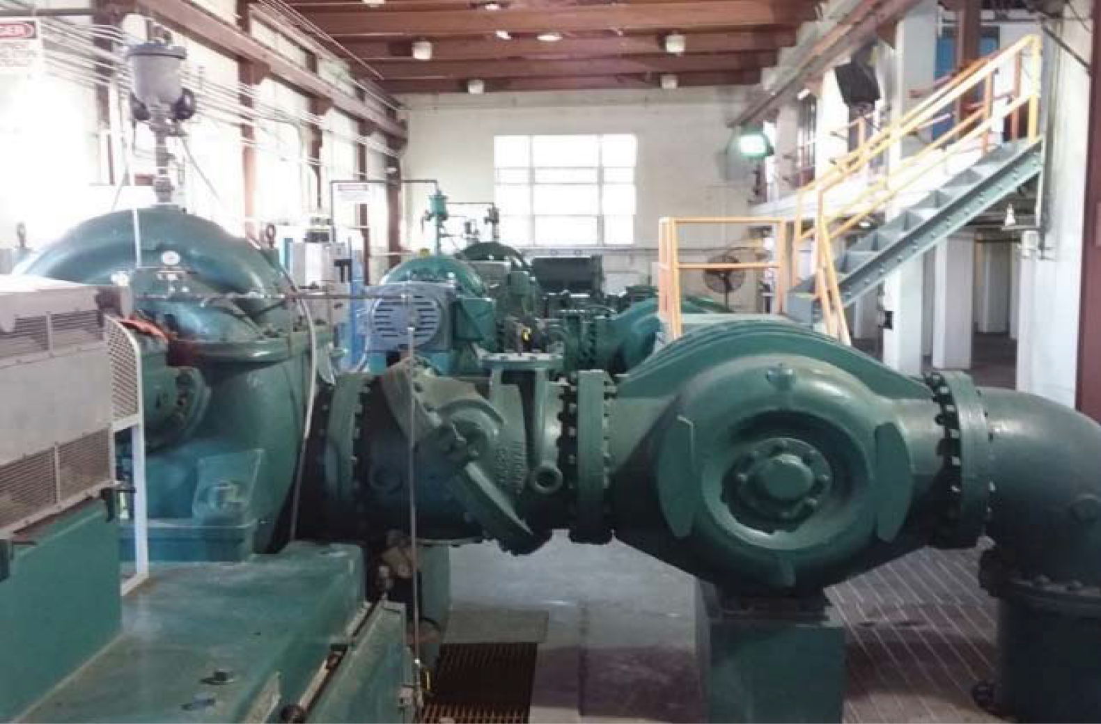 Industrial facility interior featuring large green pipes and pumps, with machinery and a staircase in the background.