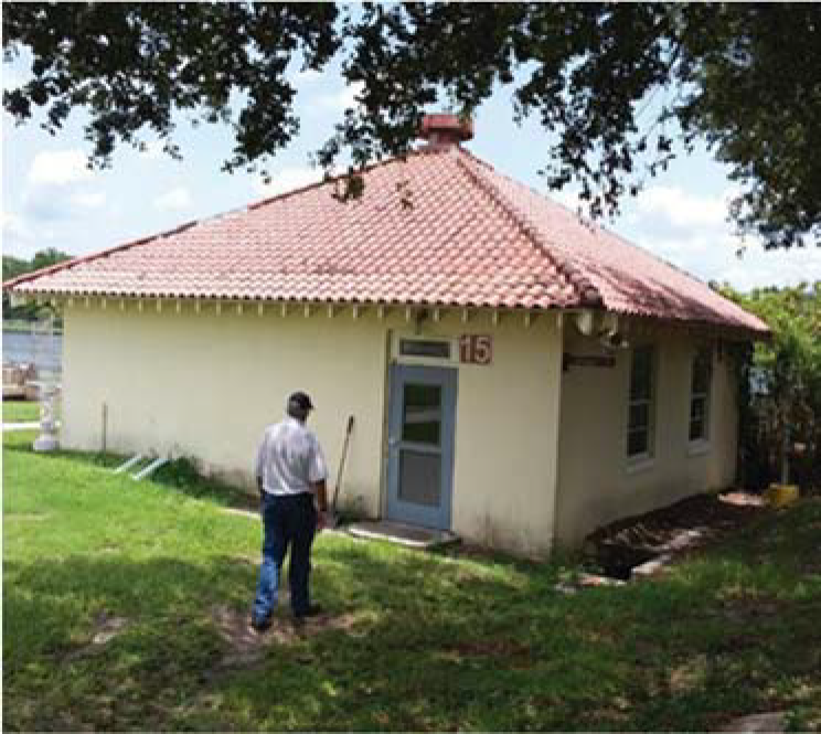 Person walking towards a small, single-story building with a red-tiled roof and numbered 15, surrounded by grass and trees.