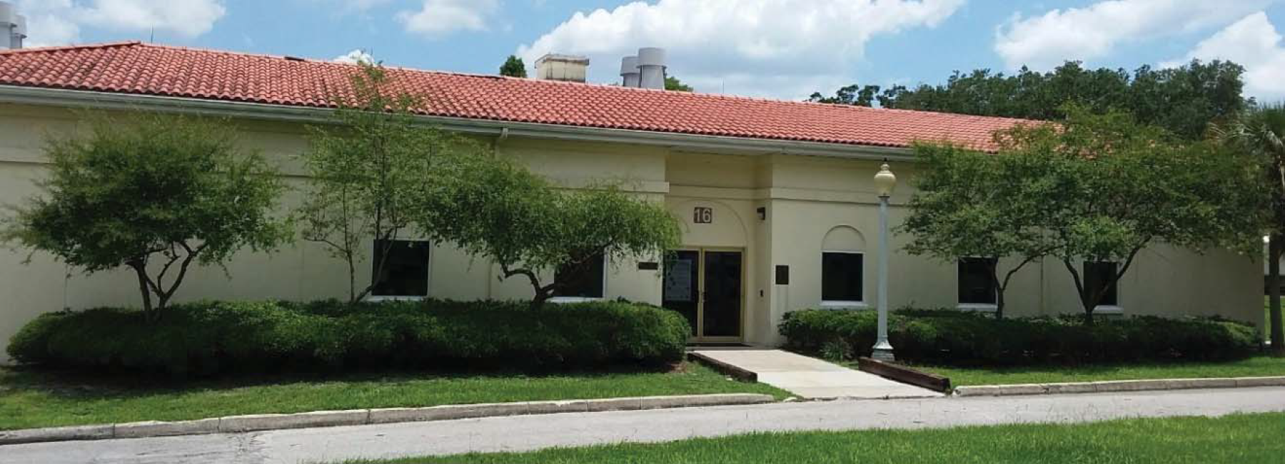 A one-story building with a red tiled roof, surrounded by green landscaping and a sidewalk in front under a blue sky.