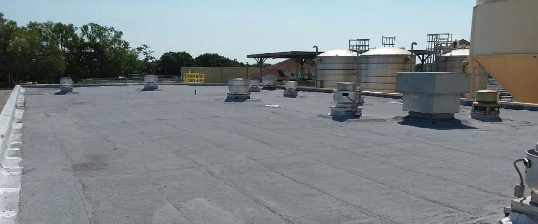 Flat rooftop with several ventilation units and large storage tanks in the background under a clear blue sky.