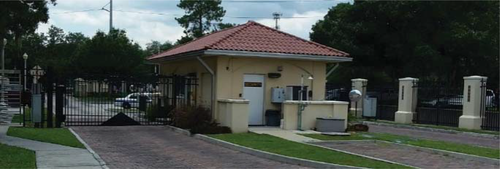 A security gatehouse with a red-tiled roof, surrounded by a fence and paved driveway, set in a green landscape.