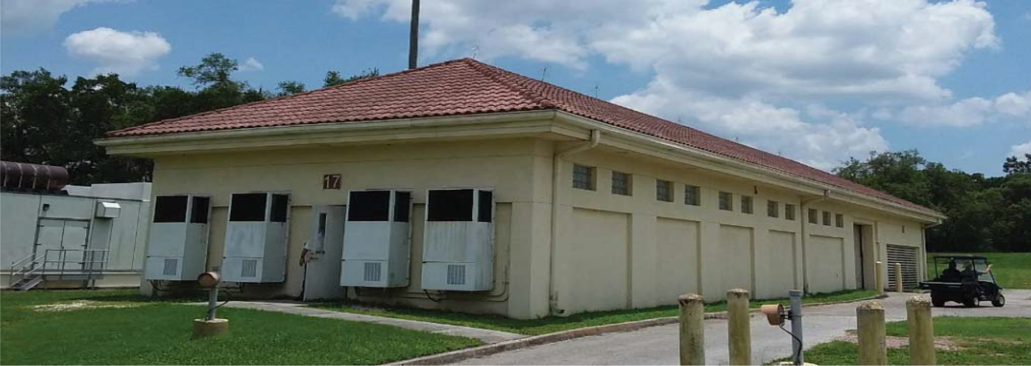 A single-story building with a red-tiled roof, multiple windows, and a grassy area, under a cloudy blue sky.