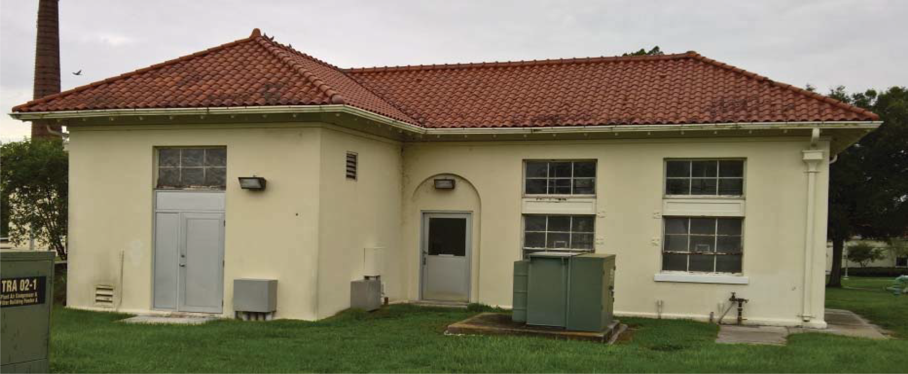 A single-story building with a red tile roof, white walls, and several windows, surrounded by green grass and utility equipment.