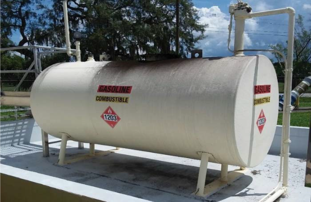 Large cylindrical gasoline storage tank with warning labels on a rooftop, under a blue sky with trees in the background.