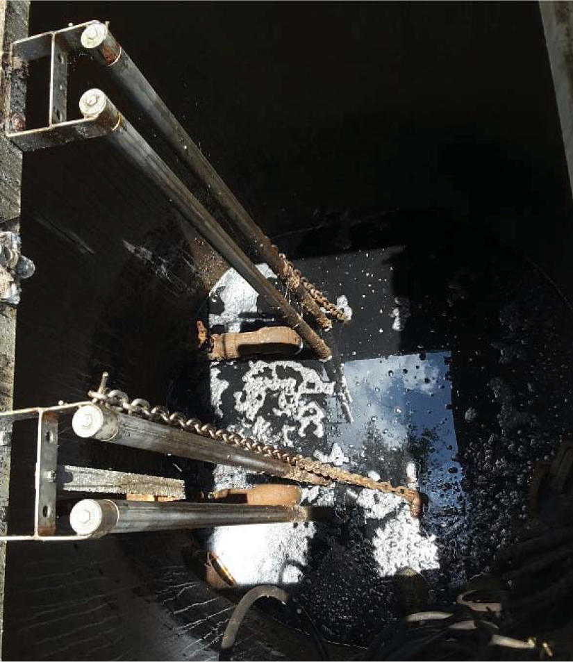 View down into a dark, cylindrical pit with chains and metal supports, surrounded by murky water and bubbles.