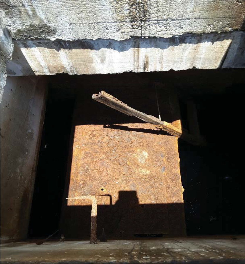 A view of a dark, rusty drain or pit with a wooden plank across it, sunlight casting shadows.