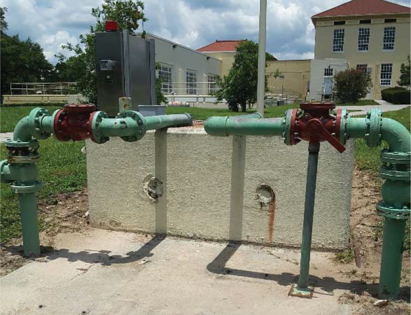 A green and red piping system with valves, situated on a concrete slab in an outdoor setting with buildings and trees in the background.