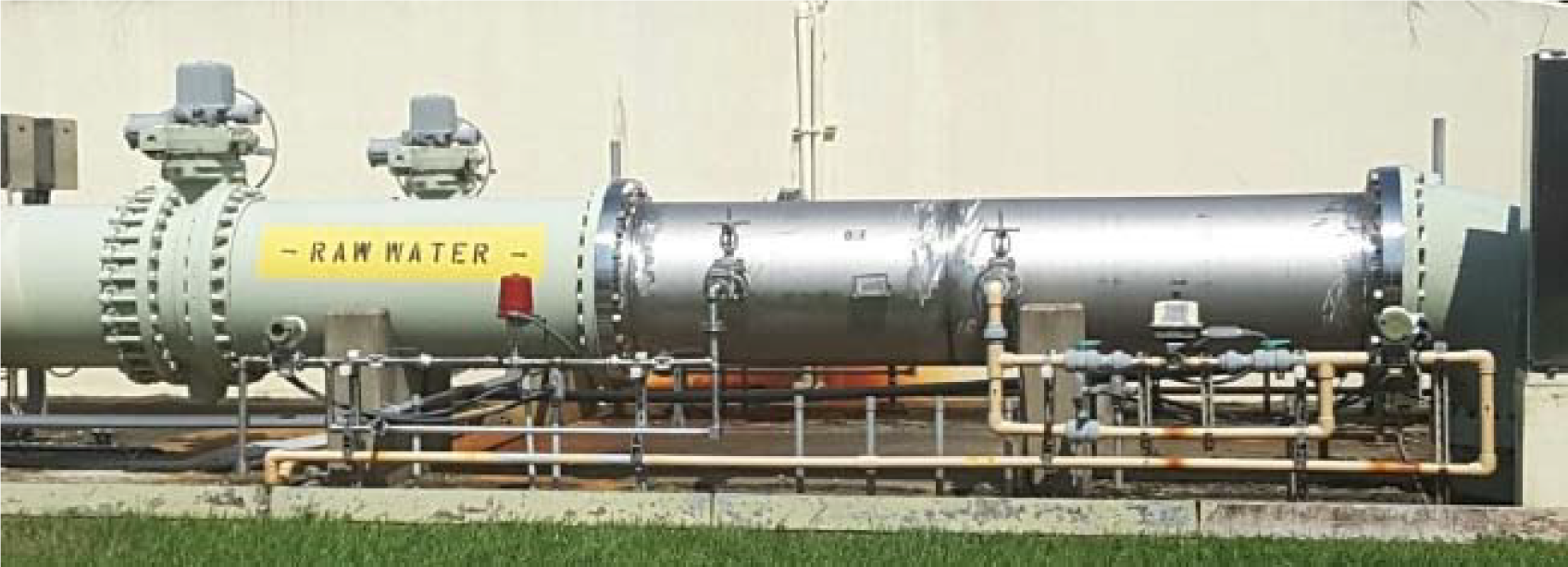 A large metal pipe labeled 'RAW WATER' with valves and connectors, situated against a wall and grass.