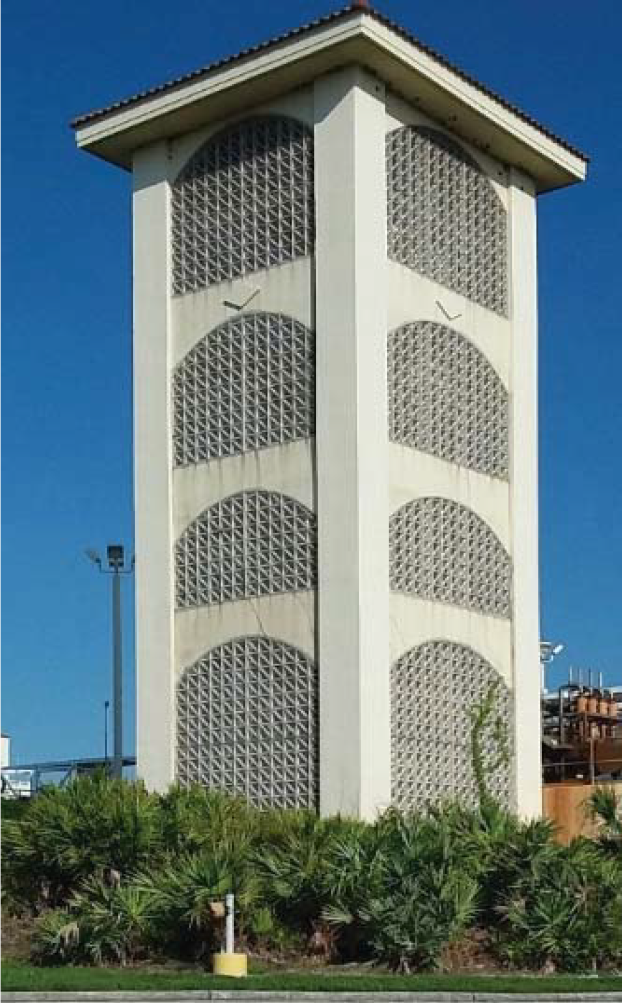 Tall white building with decorative vent-like designs, surrounded by greenery under a clear blue sky.