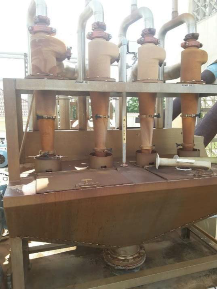 Industrial piping system with pumps and a storage tank, showing rusty components and various valves in an outdoor setting.