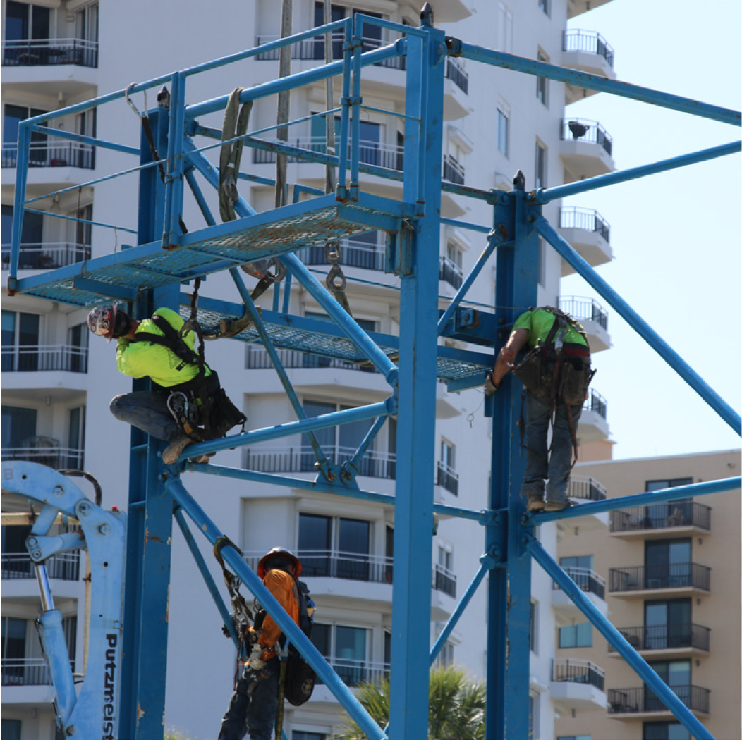 Two construction workers in safety gear climbing a blue scaffolding structure at a construction site.