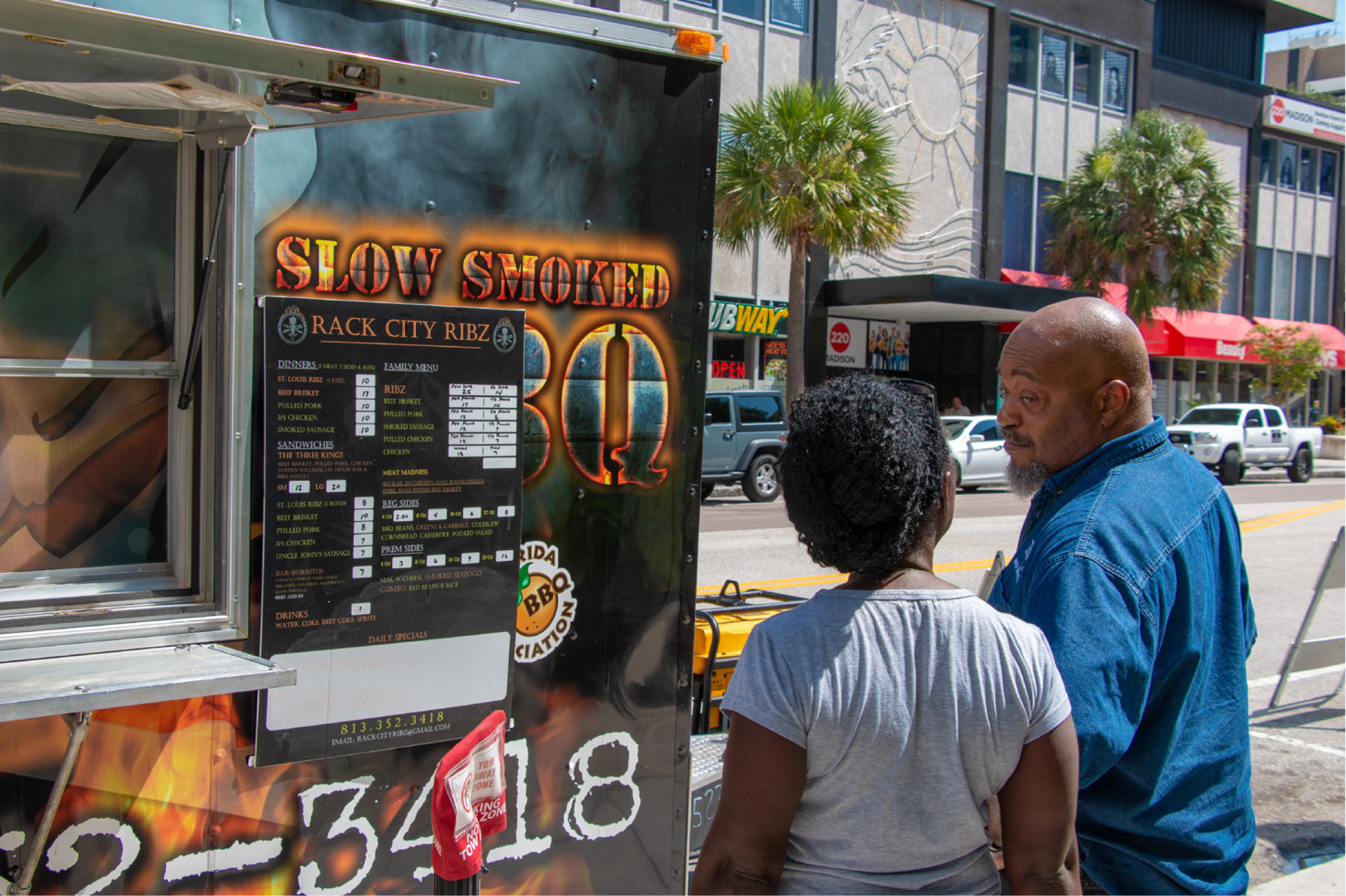 A man and a woman look at a food truck menu displaying BBQ options in an urban setting.