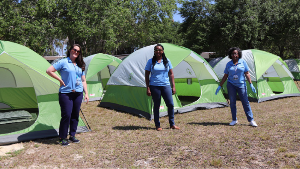 Three women in blue shirts standing beside green camping tents in a grassy area.