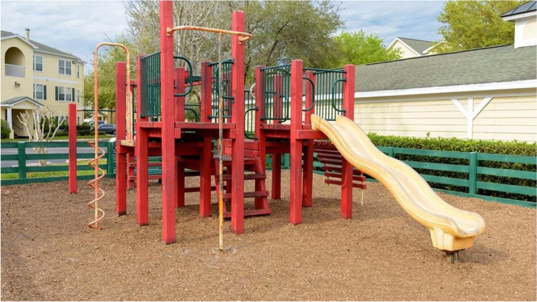 Colorful playground structure with slide, climbing stairs, and spiral climber, situated on gravel ground surrounded by green fencing and residential buildings.