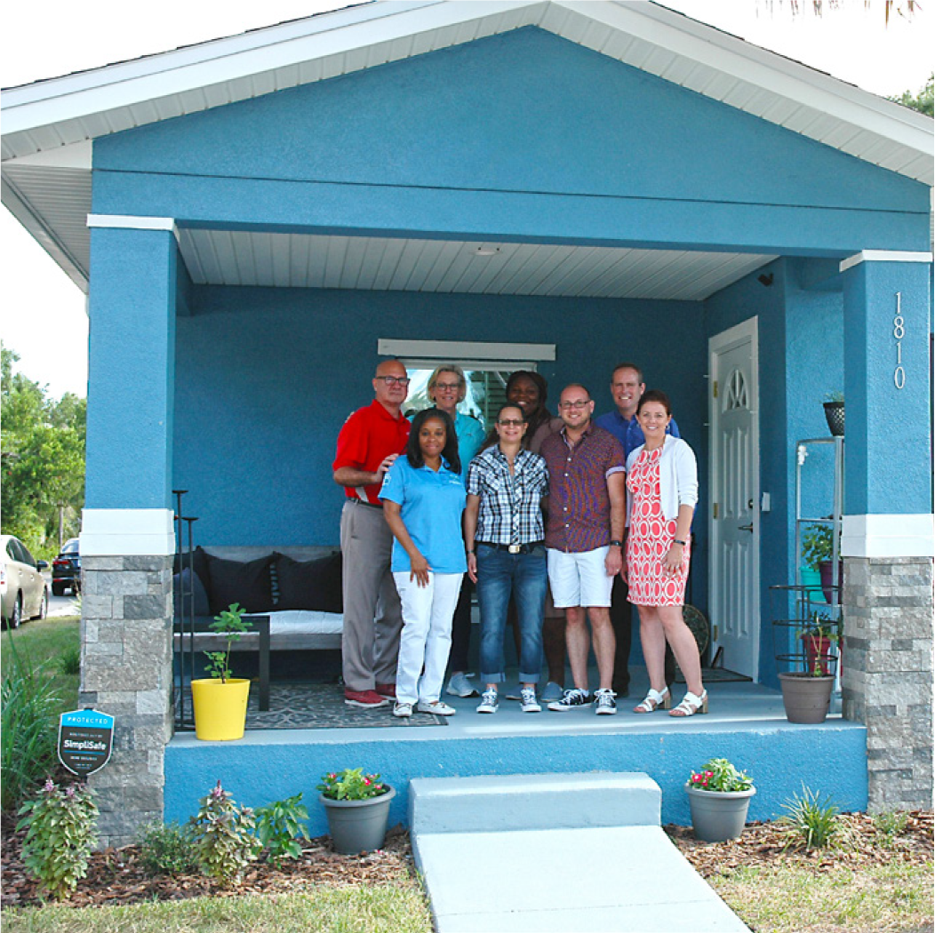 Group of eight people standing on the porch of a light blue house, smiling for the camera.
