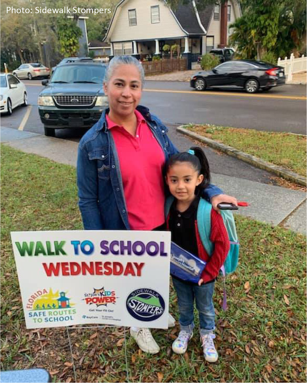 A mother and daughter stand next to a sign promoting Walk to School Wednesday, in a residential area.