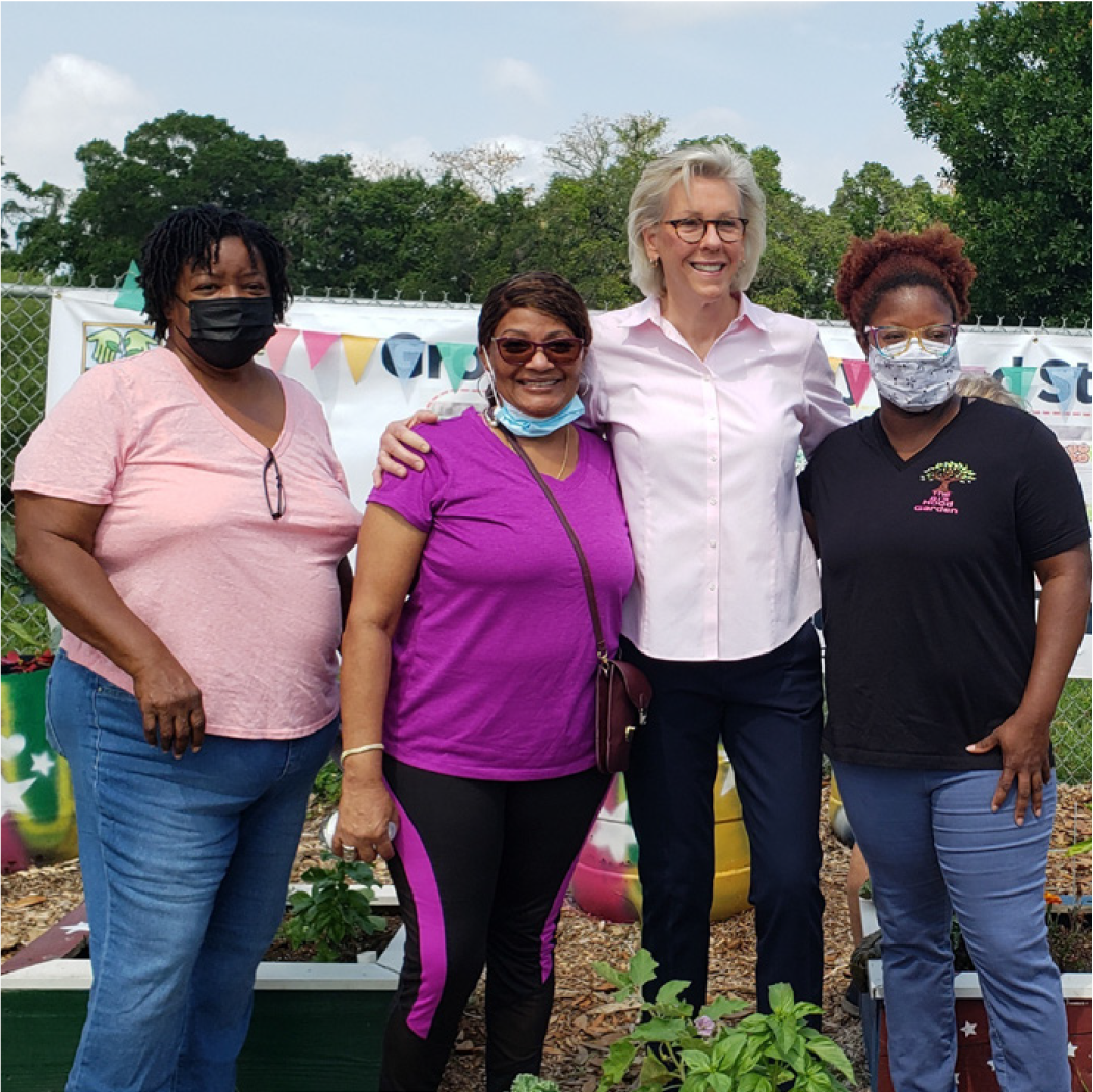 Four women smiling together in a community garden, wearing masks, with colorful bunting in the background.