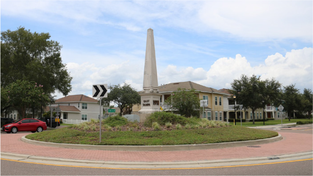 A roundabout featuring an obelisk monument surrounded by greenery and residential buildings under a blue sky with clouds.