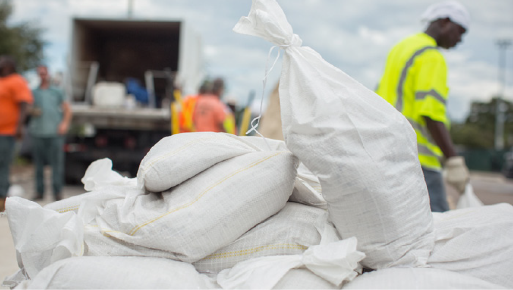 White sacks stacked, with workers in safety gear and a truck in the background, indicating a loading or delivery activity.