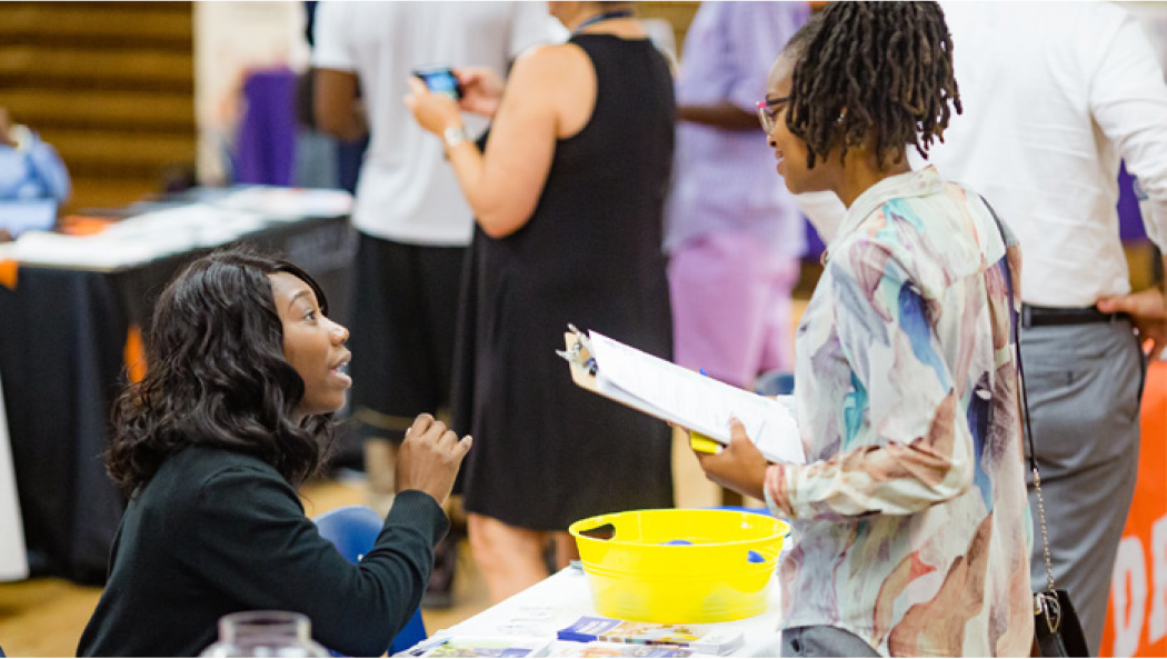 A woman converses with another woman while holding a clipboard in a busy event setting.