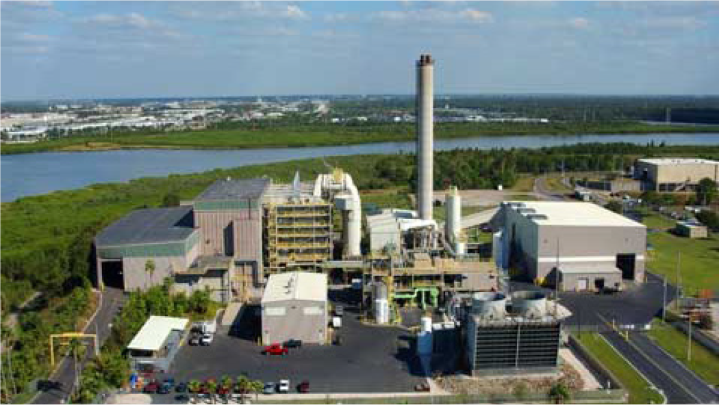 Aerial view of an industrial facility near a river, featuring buildings, a tall smokestack, and vehicles in the parking area.