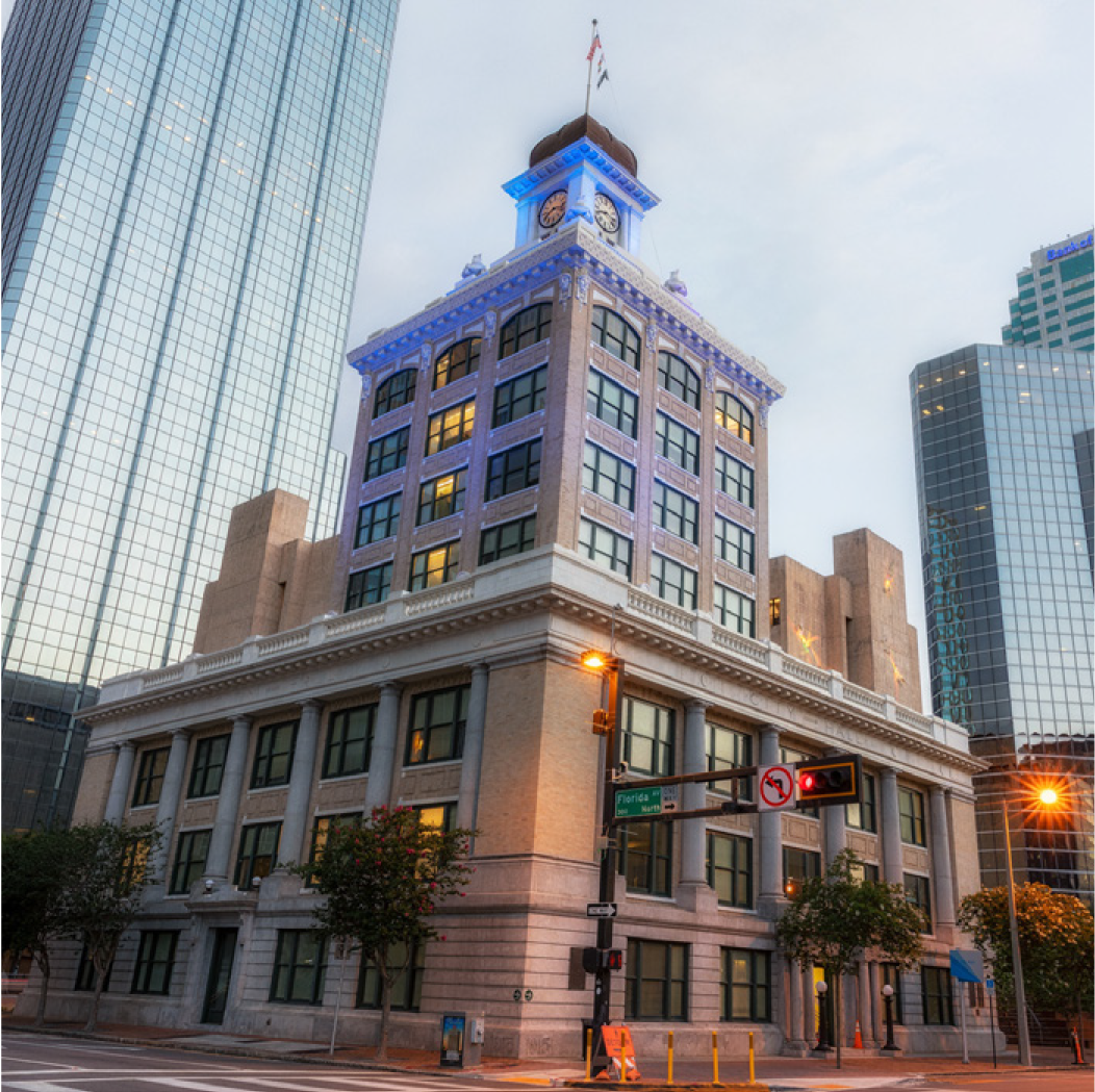 Historic building with a clock tower illuminated in blue, surrounded by modern skyscrapers and streetlights.