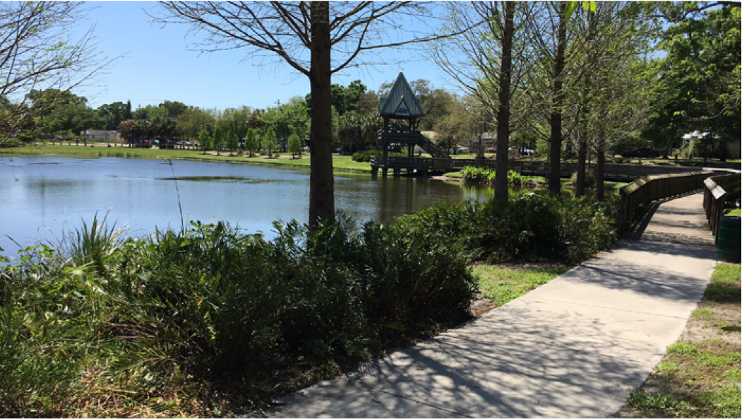 A serene park scene with a pathway along a lake, trees, and a gazebo in the background under a clear blue sky.
