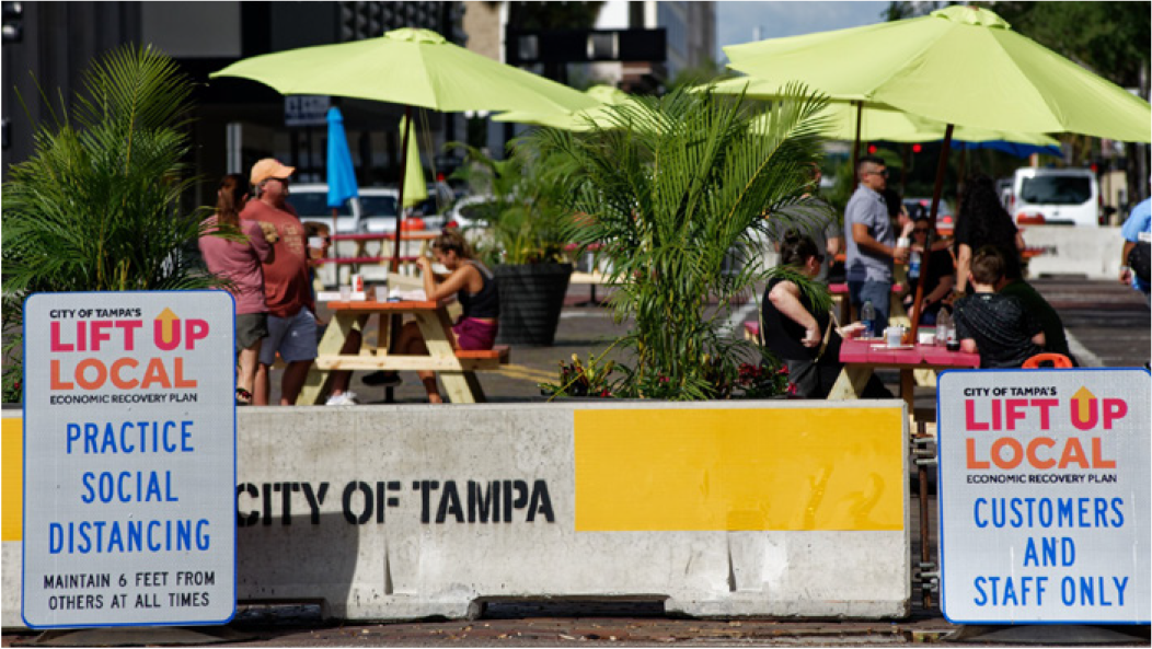 Outdoor dining area with green umbrellas, signage promoting social distancing, and people enjoying meals in Tampa.