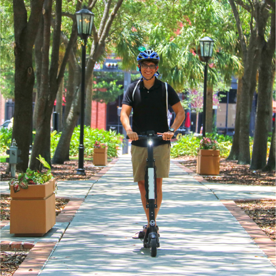A smiling person wearing a helmet rides an electric scooter down a tree-lined pathway with planters on either side.