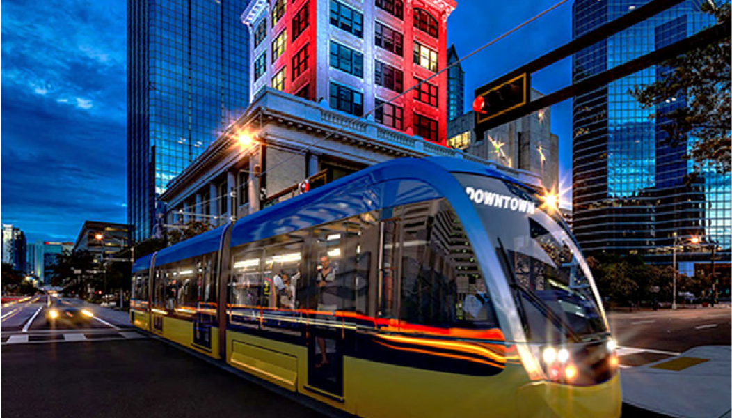 A blue and yellow streetcar in downtown, with a brightly lit building and skyscrapers in the background during twilight.