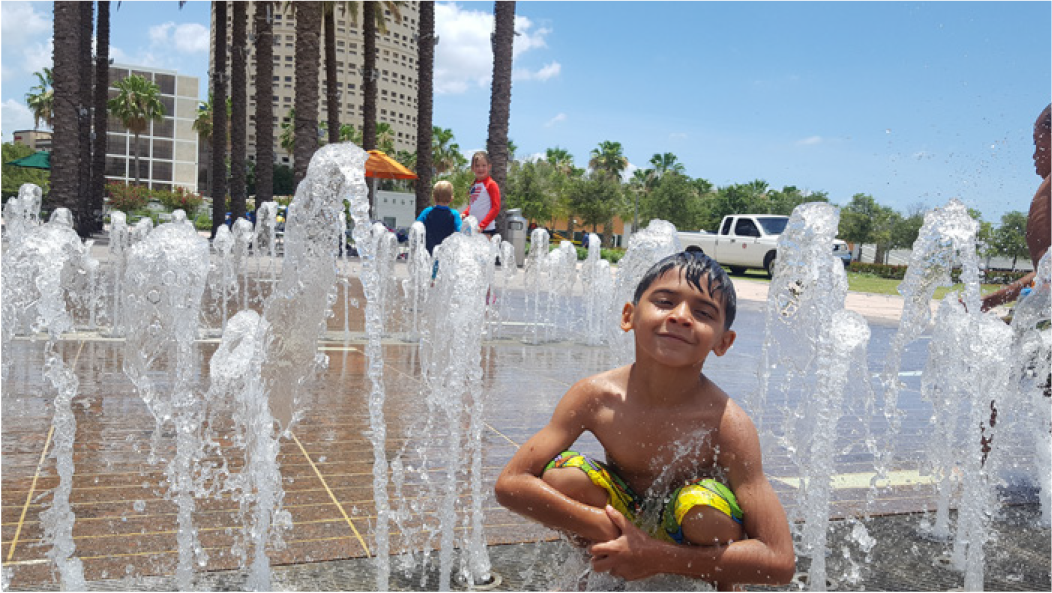 A joyful boy crouches in a splash pad, surrounded by water jets, with people playing in the background on a sunny day.