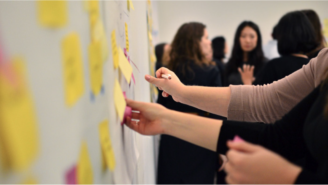 A group of people collaborating around a wall covered in sticky notes, with hands actively placing and adjusting the notes.