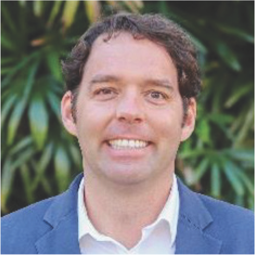 A smiling man with curly hair wearing a blue blazer and white shirt, standing outdoors among leafy green plants.