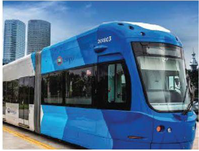 A blue tram stopped on a city street with skyscrapers in the background and clear sky.