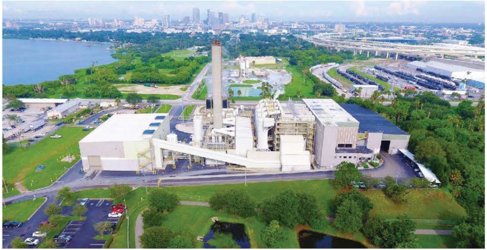 Aerial view of an industrial facility near a body of water and city skyline, surrounded by green landscapes and roads.