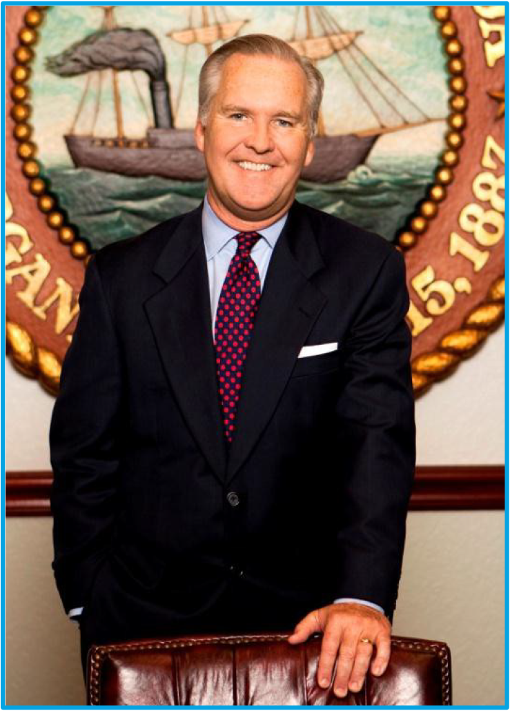 Smiling man in a suit stands confidently in front of a nautical-themed emblem and a wooden chair.