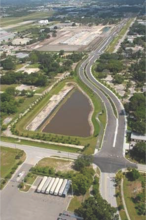 Aerial view of a roadway and a water body surrounded by greenery and industrial buildings in the background.