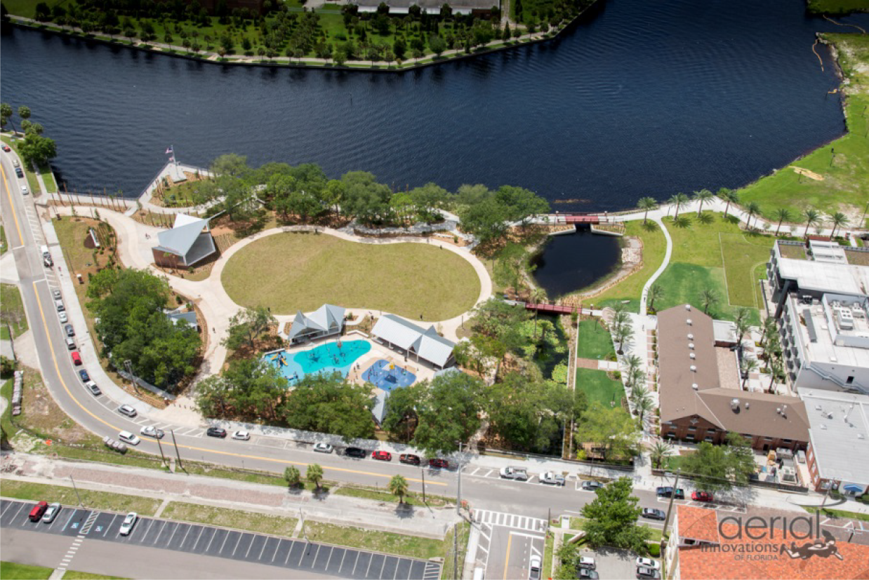 Aerial view of a park by a river, featuring a pond, swimming pool, and green spaces.