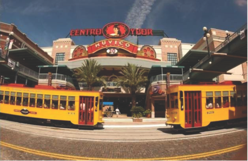 Two yellow streetcars in front of the Centro Ybor shopping center, with a blue sky and palm trees in the background.