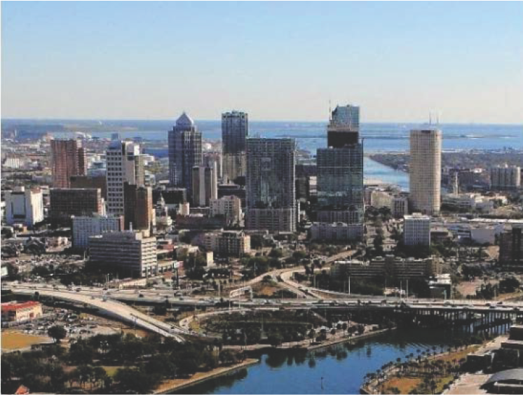 Aerial view of a city skyline featuring tall buildings, a river, and green spaces under a clear blue sky.