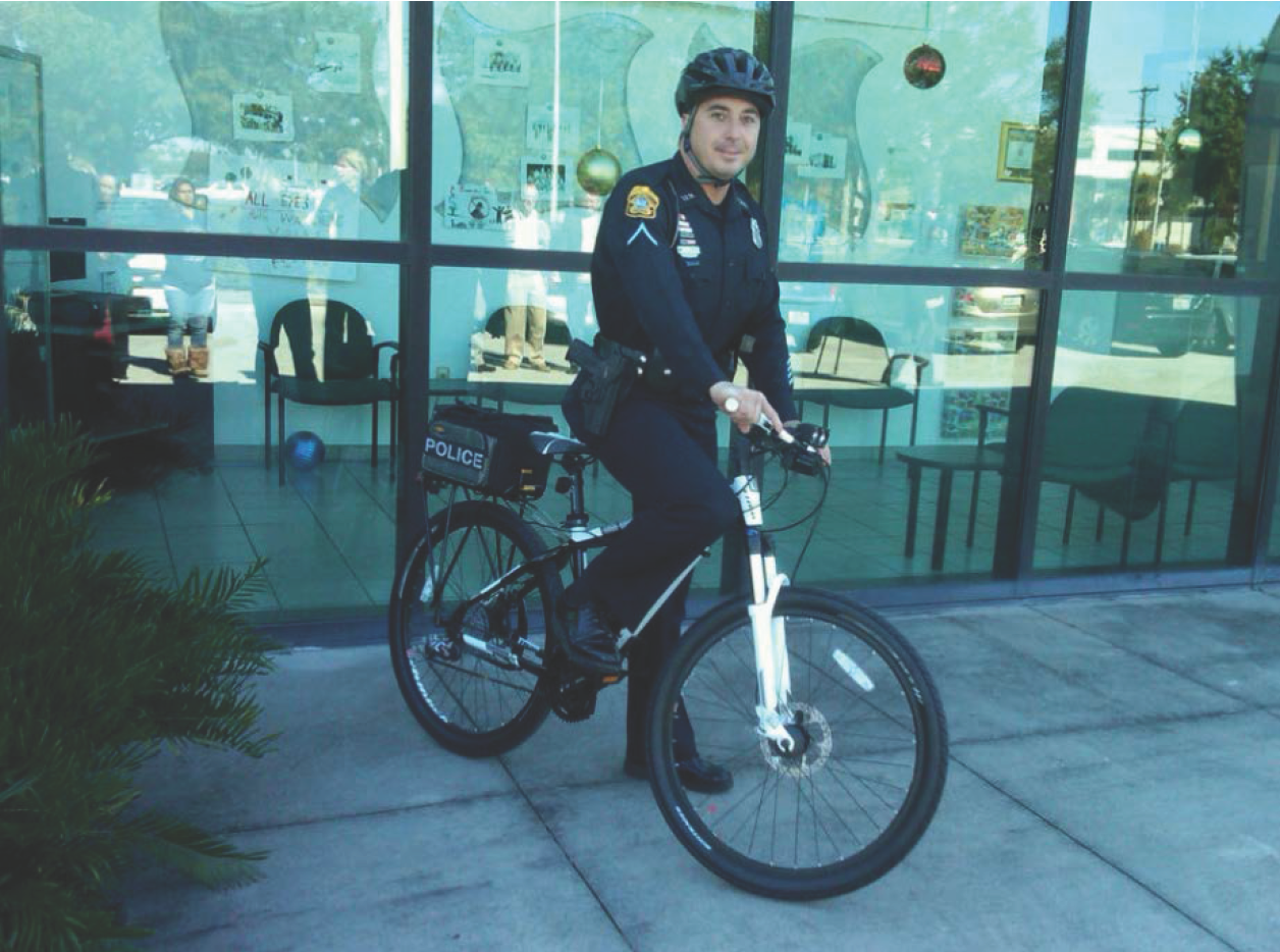 A police officer in uniform sits on a bicycle outside a building, with seated visitors visible through the glass.