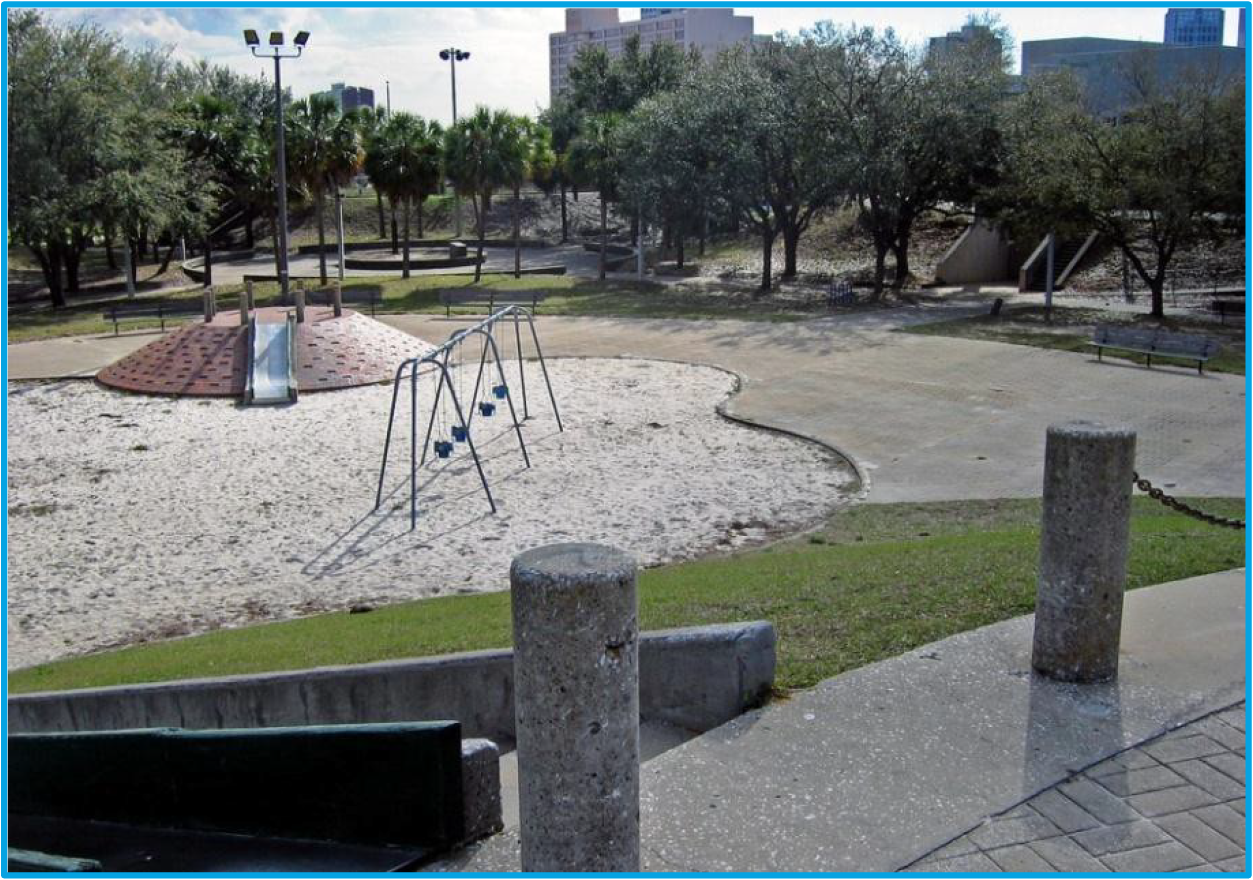 A playground with swings and a climbing structure, surrounded by sandy areas and trees in an urban park setting.