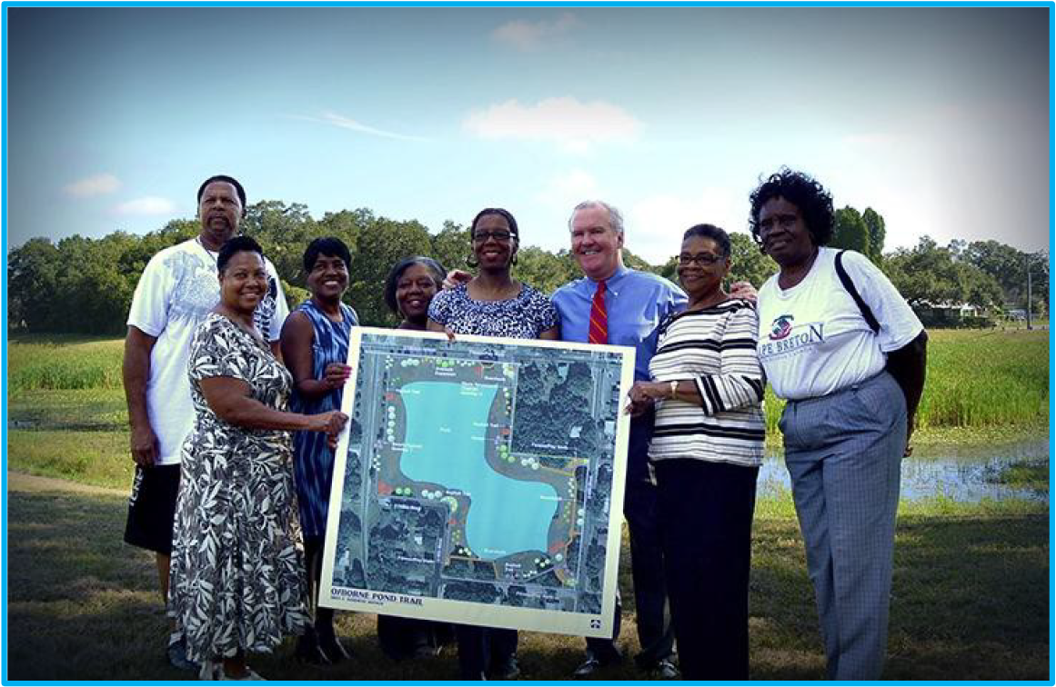 Group of people holding a map by a lake, smiling under a blue sky.