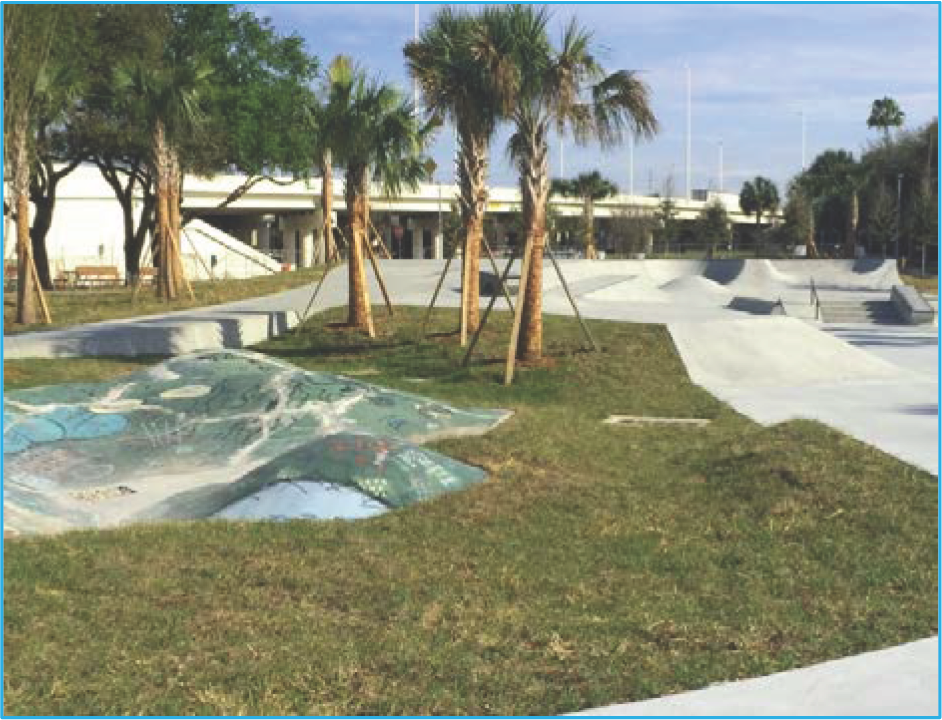 A skate park with various ramps and a grassy area, surrounded by palm trees under a clear sky.