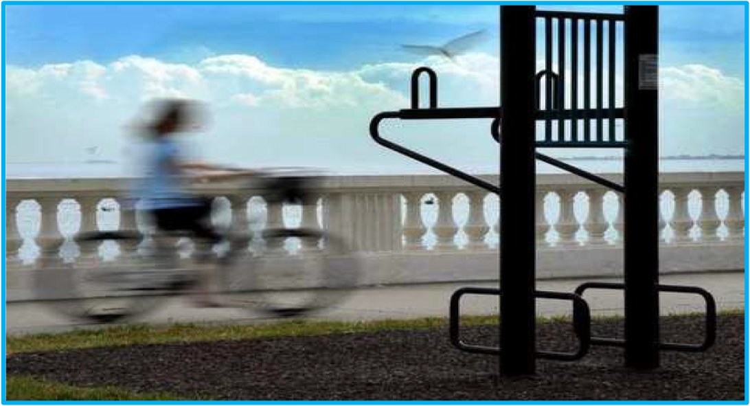 A blurred cyclist rides past an outdoor exercise station along a waterfront path with blue skies and clouds in the background.
