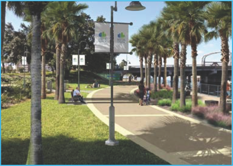 A scenic waterfront park featuring palm trees, walking paths, benches, and banners, with a bridge in the background under a clear blue sky.