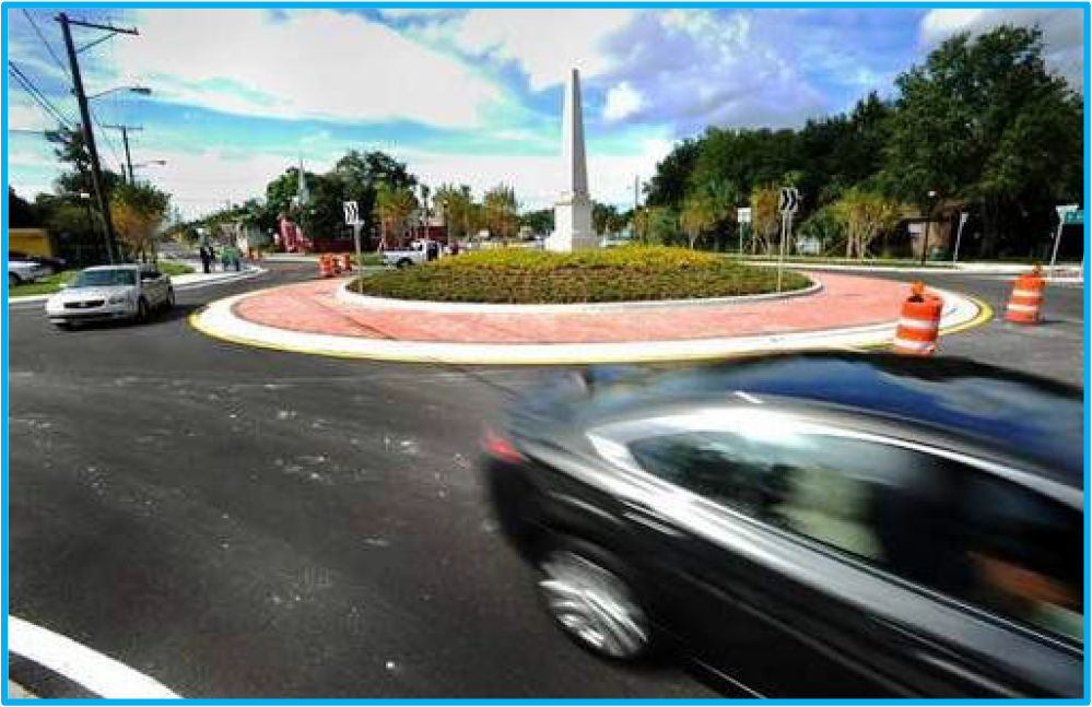 A busy roundabout with cars navigating, an obelisk in the center and construction cones visible.