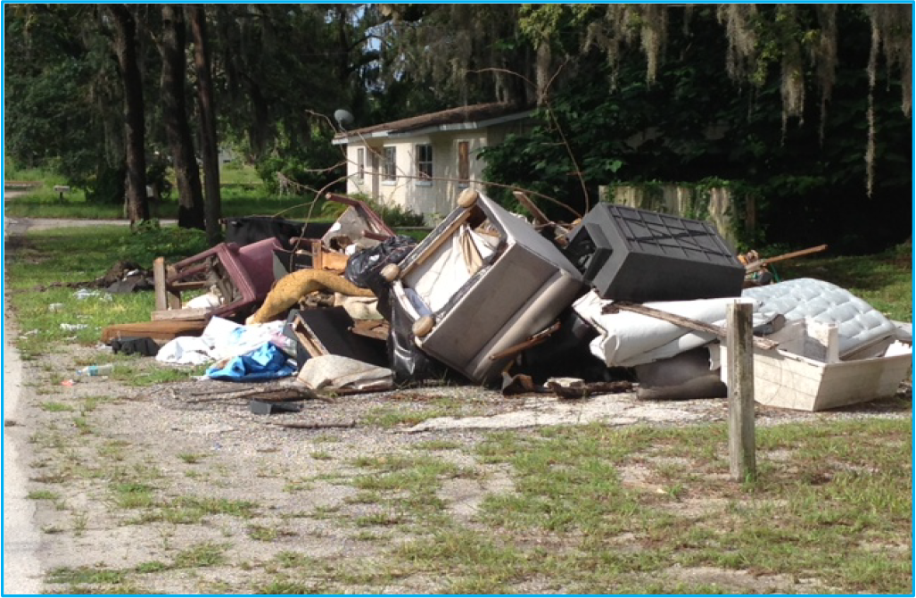 A pile of discarded furniture and debris beside a roadway, with trees and a house in the background.