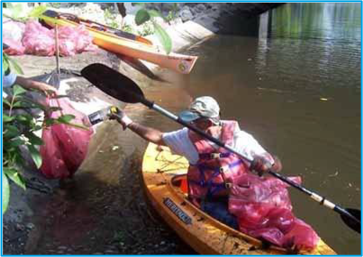 A kayaker wearing a life jacket retrieves a bag of trash from the water near a bridge, promoting environmental cleanup.