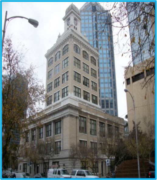 Historic building with clock tower in an urban setting, surrounded by modern skyscrapers and trees.